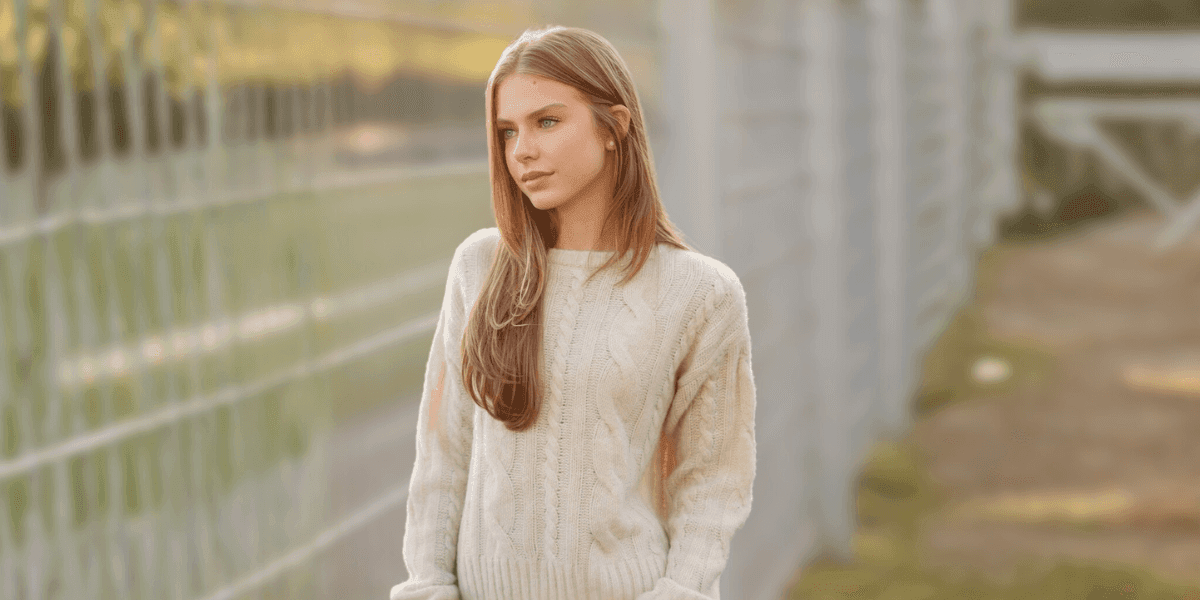 "Young woman in a beige sweater, standing against a blurred glass background, exuding a calm and introspective presence."