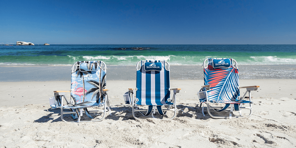 Three beach chairs in different vibrant patterns, placed on a sandy beach with the ocean waves in the background, perfect for relaxation under the sun.