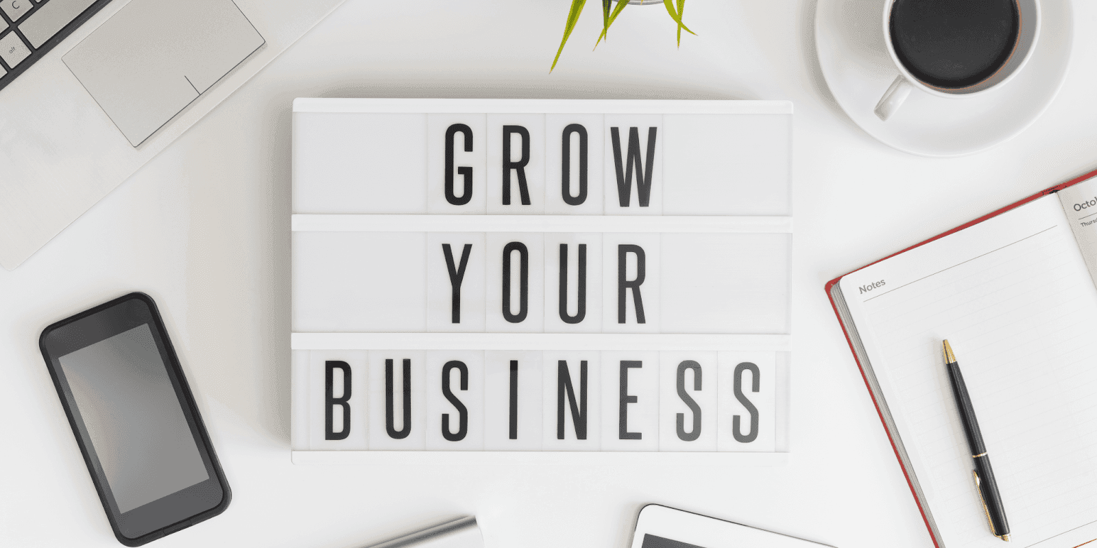 Workspace with laptop, phone, coffee, and notebook surrounding lightbox sign reading “Grow Your Business”.