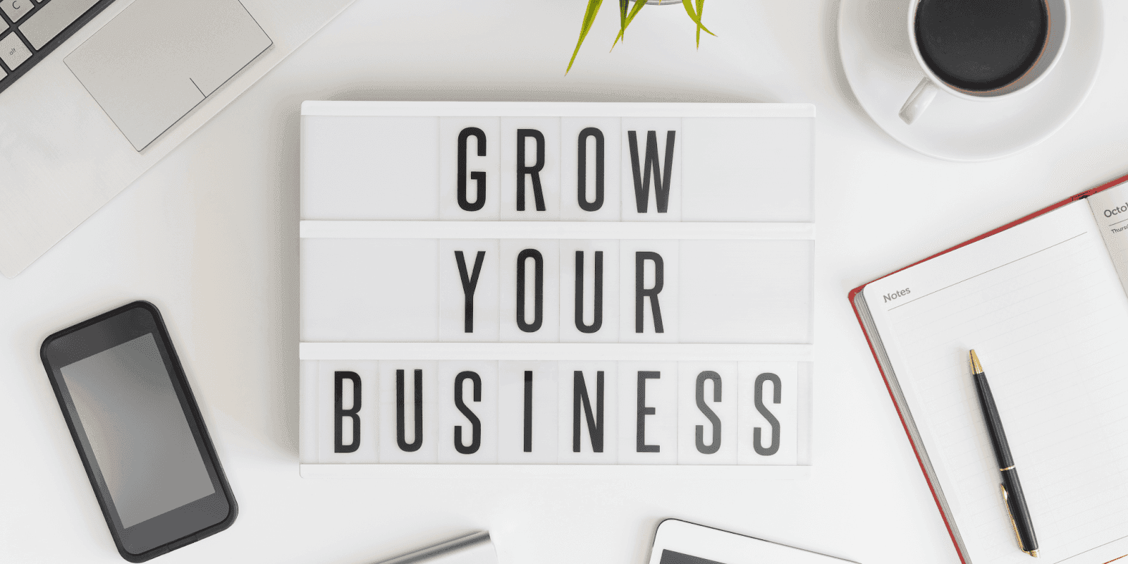 Workspace with laptop, phone, coffee, and notebook surrounding lightbox sign reading “Grow Your Business”.