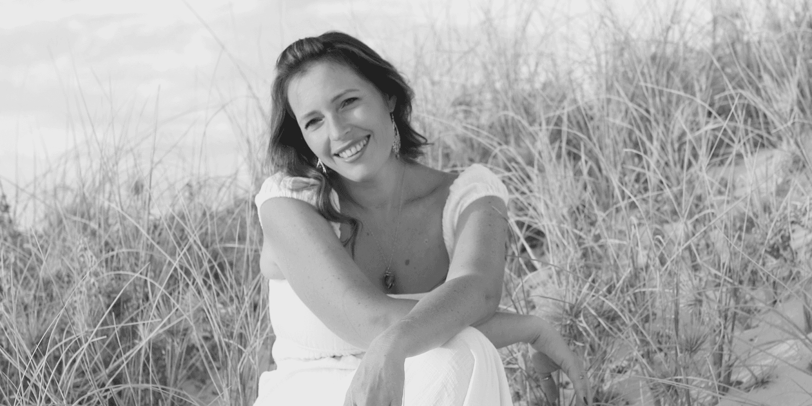 Smiling woman sitting on the sand at the beach, wearing a white dress and earrings, in black and white photo with tall grass in the background.