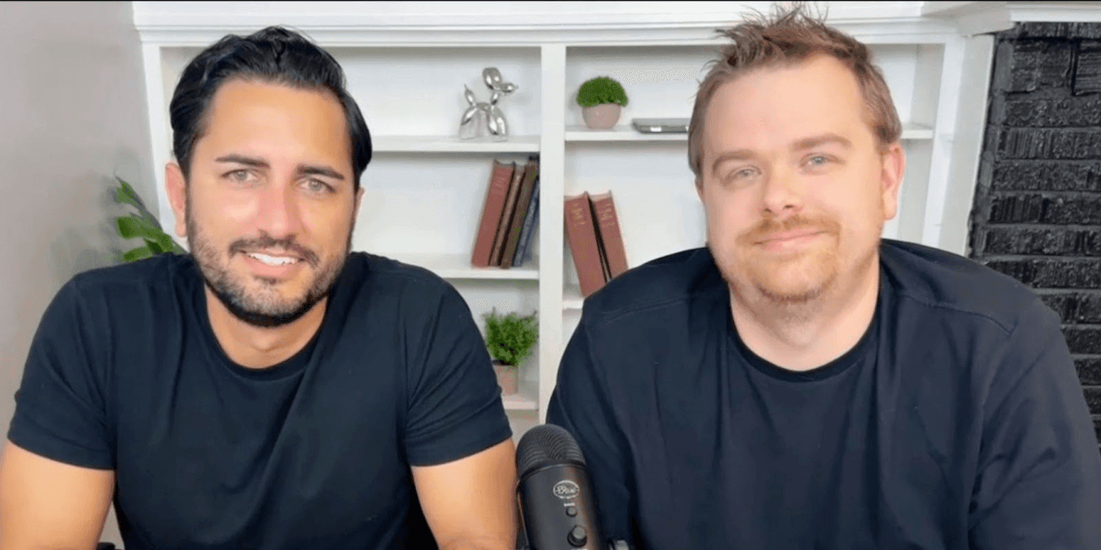 "Two men smiling at the camera while sitting at a table in a well-lit space, with bookshelves and microphones in the background."