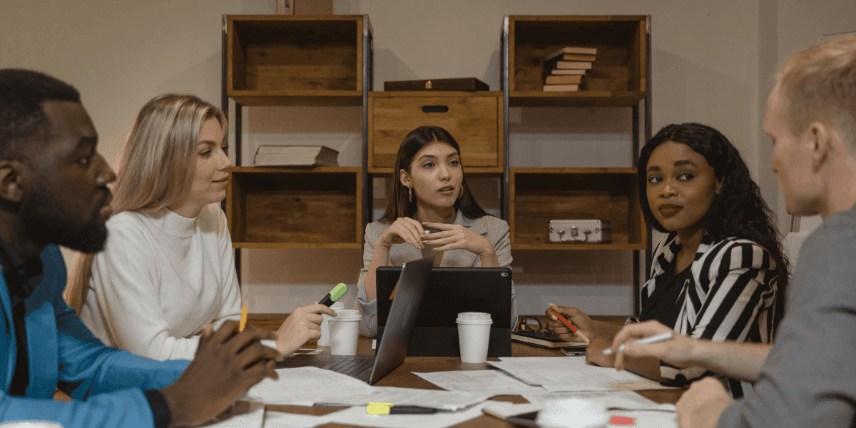 A diverse group of five professionals engaged in a meeting around a conference table. They are discussing ideas with laptops, notepads, and coffee cups in front of them. The individuals are focused on the conversation, with one person speaking and the others listening attentively. The background features wooden shelves with books, adding to the professional ambiance of the meeting space.