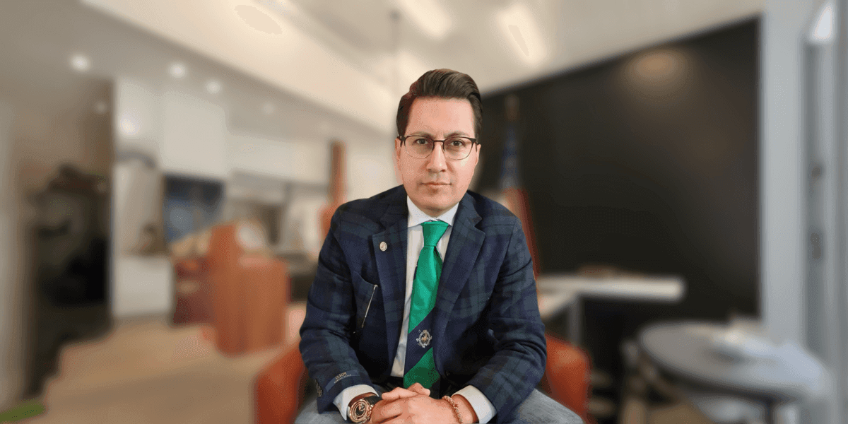 "Professional man dressed in a plaid suit and green tie, sitting with hands folded in front of him, exuding confidence in a modern office setting with a blurred background."