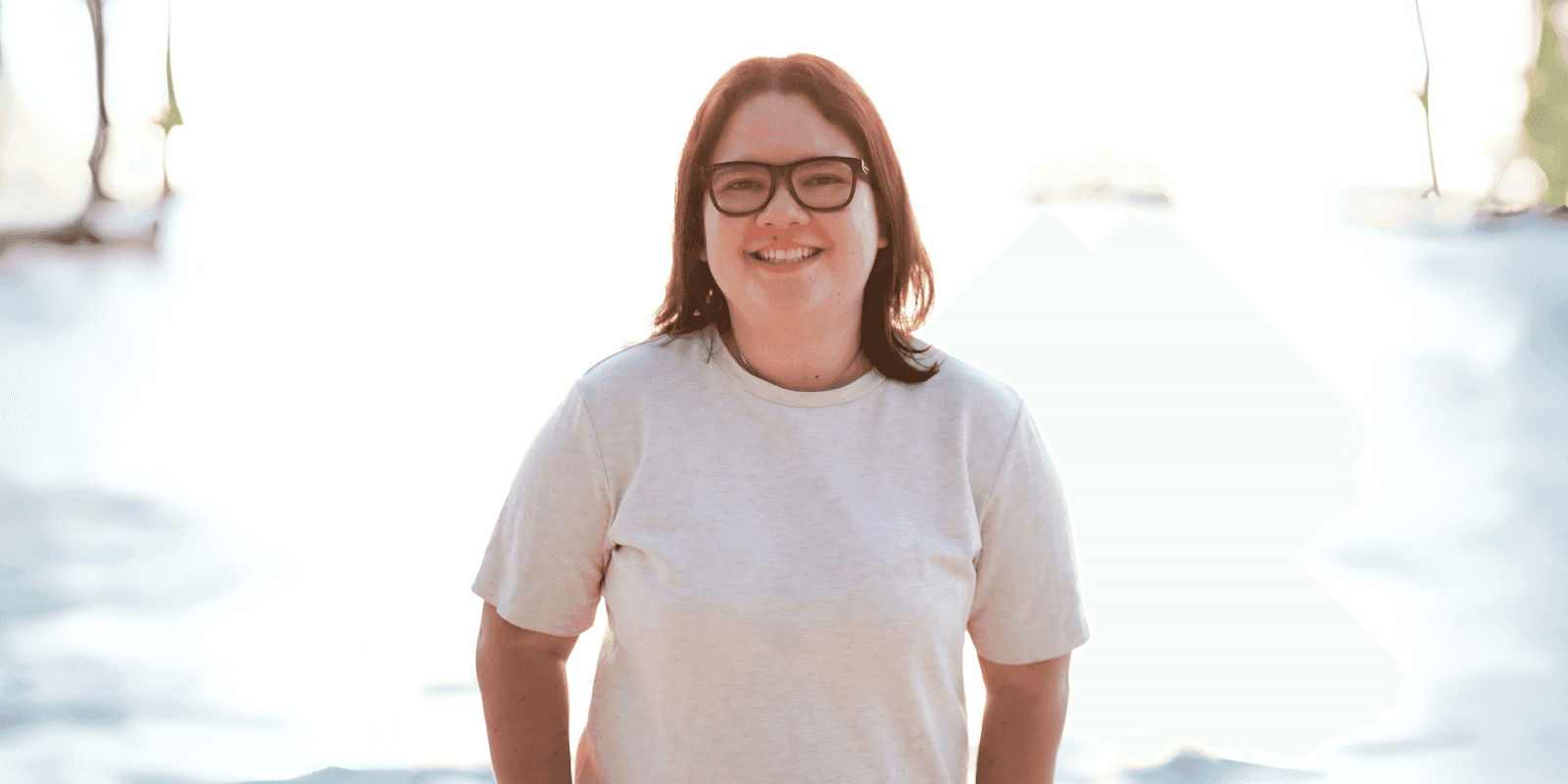 "Smiling woman in a neutral-toned shirt with short hair, standing outdoors with bright sunlight in the background, exuding a cheerful and relaxed vibe."
