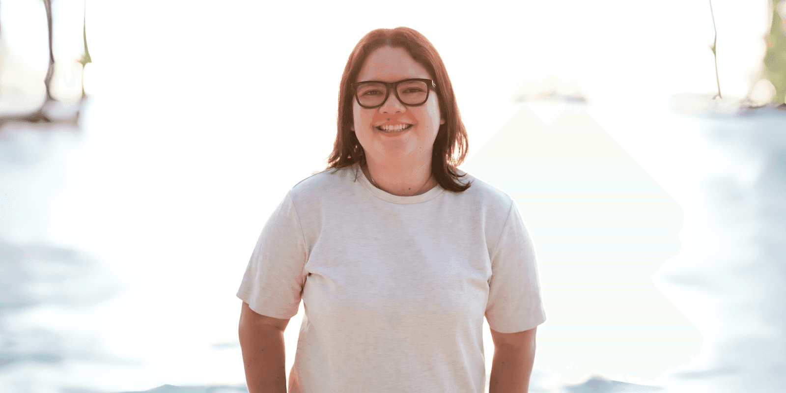 "Smiling woman in a neutral-toned shirt with short hair, standing outdoors with bright sunlight in the background, exuding a cheerful and relaxed vibe."