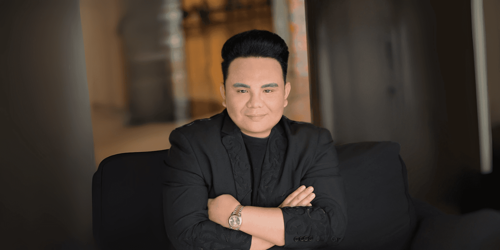 Tommie Mopia sits confidently in a black suit with arms crossed, smiling at the camera.