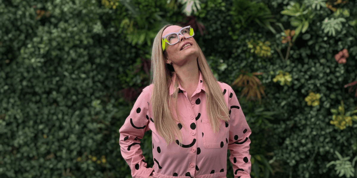 Emmaline McAndrew, founder of The Slow Businesswoman, smiling in a polka-dot shirt and colorful glasses, standing against a green background.