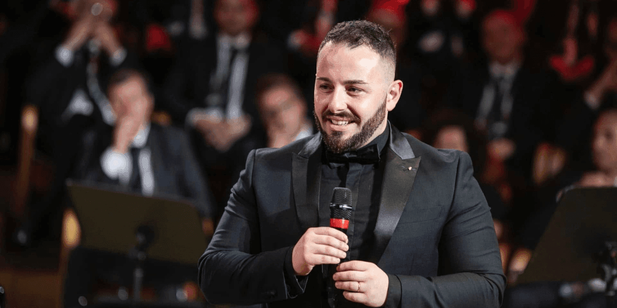 Salvatore Frega smiling while holding a microphone during a performance at a formal event with an orchestra in the background.