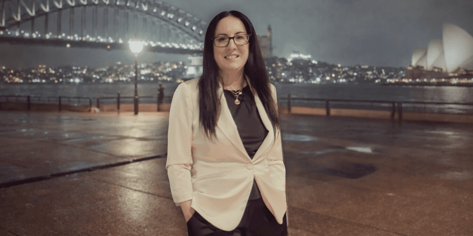 Woman smiling by Sydney Harbour Bridge at night with city lights and Opera House in background.