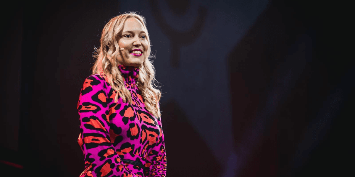 Simone Herzog speaking onstage in a colorful patterned outfit with a headset microphone.