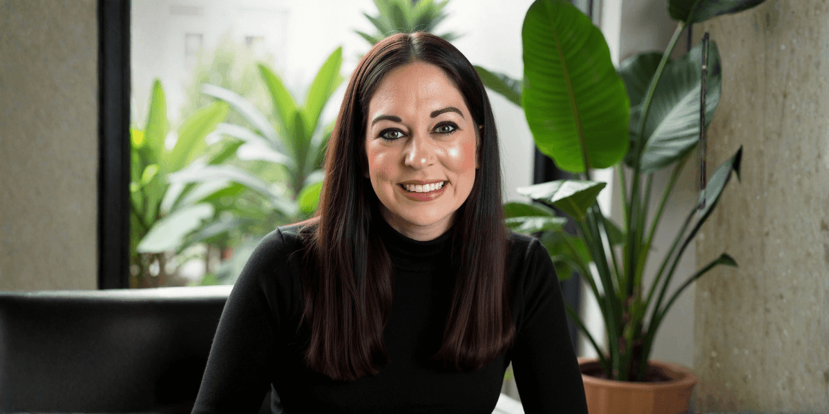 Author Jessica Brenner smiling in a black turtleneck, seated indoors with large green plants in the background.