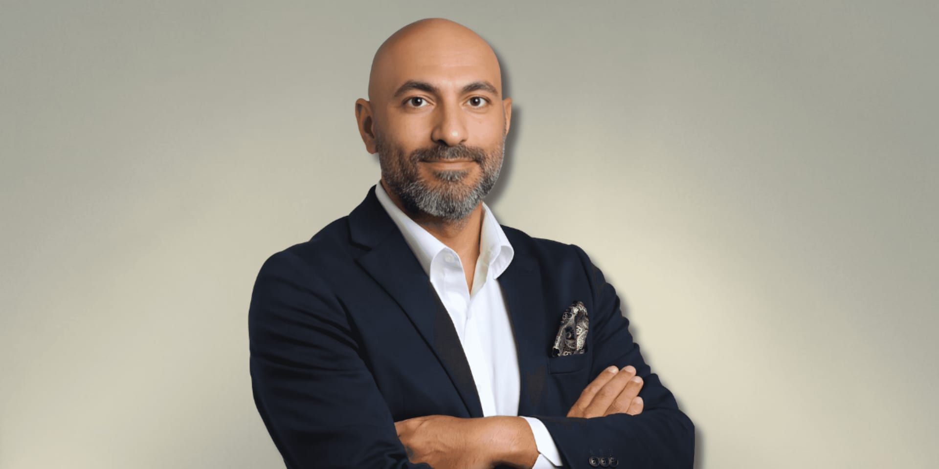 A man with a short beard and a bald head is dressed in a navy blue suit with a white shirt and a patterned pocket square. He is standing with his arms crossed and smiling, against a soft neutral-colored background.