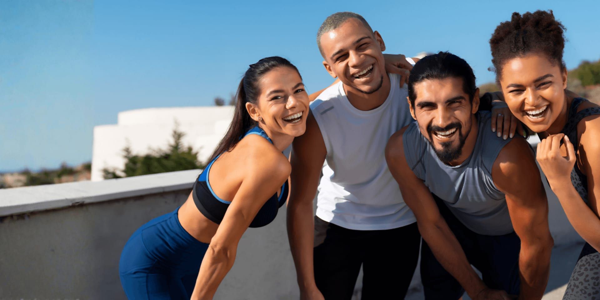 A group of four people, two men and two women, smiling and leaning close together in athletic wear. They appear joyful against a clear blue sky background.
