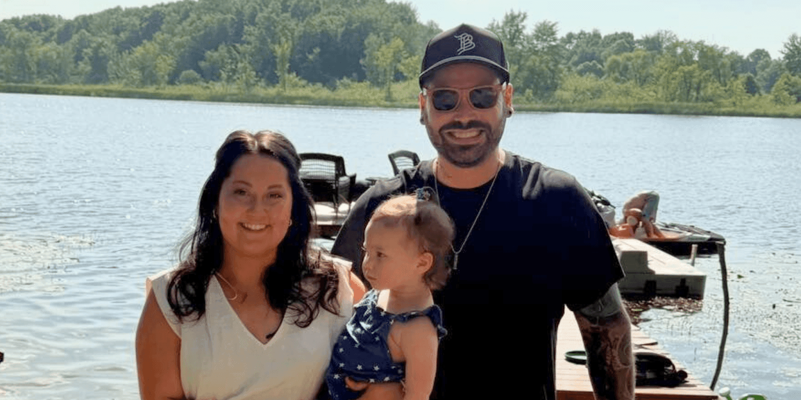 Family posing by the lake on a sunny day, with a man, woman, and toddler enjoying the outdoors
