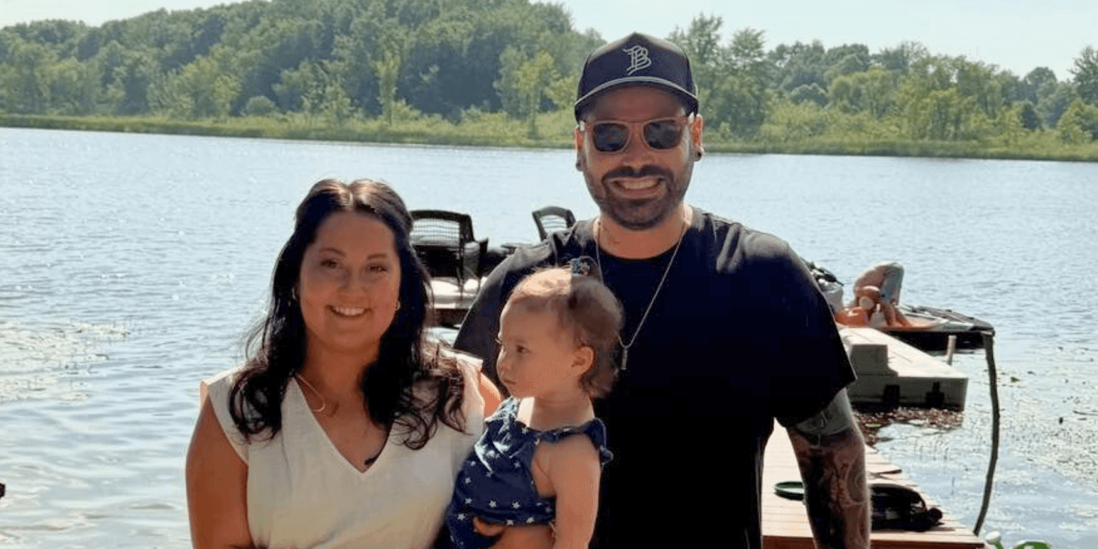 Family posing by the lake on a sunny day, with a man, woman, and toddler enjoying the outdoors