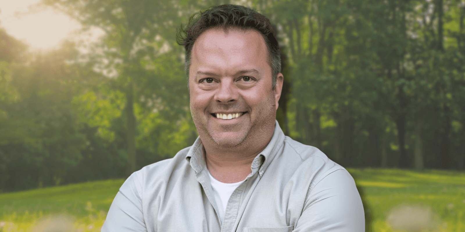 Smiling man standing in a lush, green forest with sunlight shining through trees, wearing a casual beige shirt.