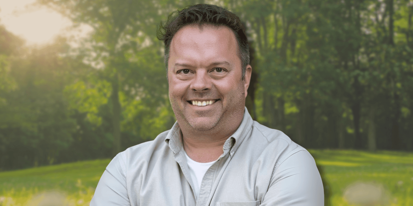 Smiling man standing in a lush, green forest with sunlight shining through trees, wearing a casual beige shirt.