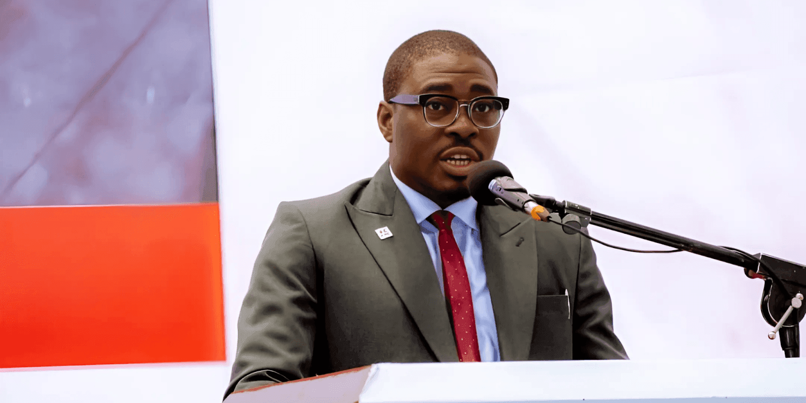 "A man in a gray suit with a red tie speaking into a microphone at a podium during an event, conveying a confident and engaging presence."