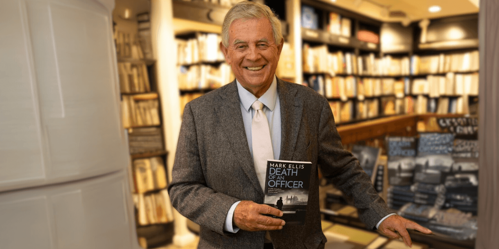 "Mark Ellis holding a copy of his book 'Death of an Officer' in a bookstore setting, smiling and dressed in a suit, showcasing the author and his work."
