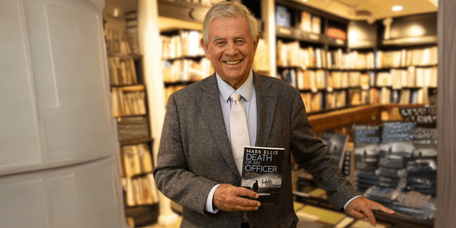 "Mark Ellis holding a copy of his book 'Death of an Officer' in a bookstore setting, smiling and dressed in a suit, showcasing the author and his work."