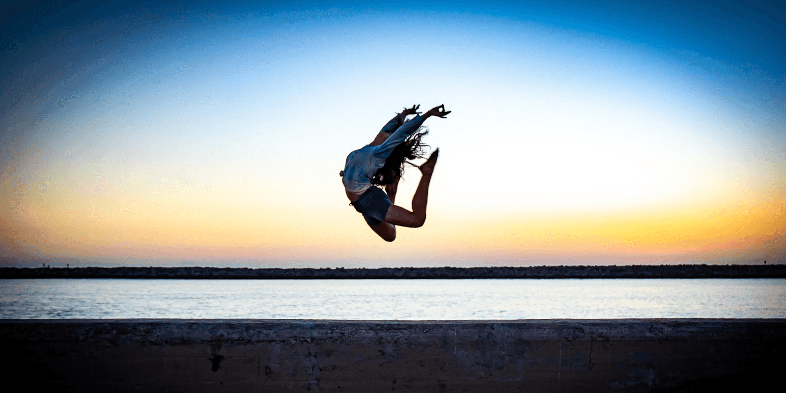 A dancer gracefully leaps into the air by the water, captured in a vibrant sunset setting, symbolizing joy and freedom.