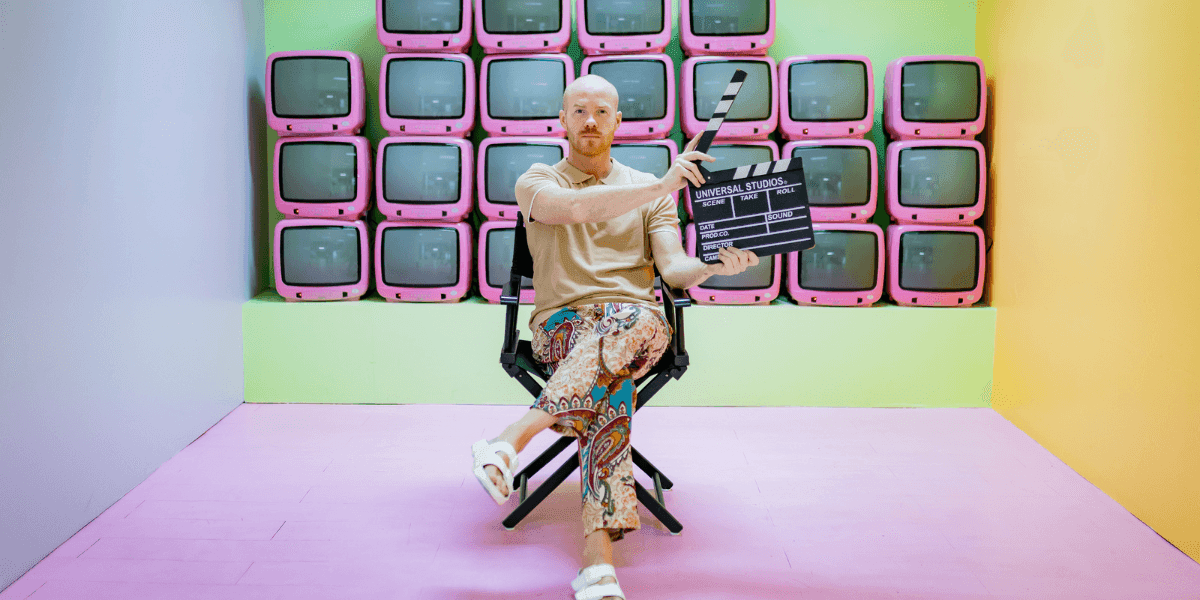 Thom Rikko holds a film clapperboard while seated before a colorful wall of vintage televisions.