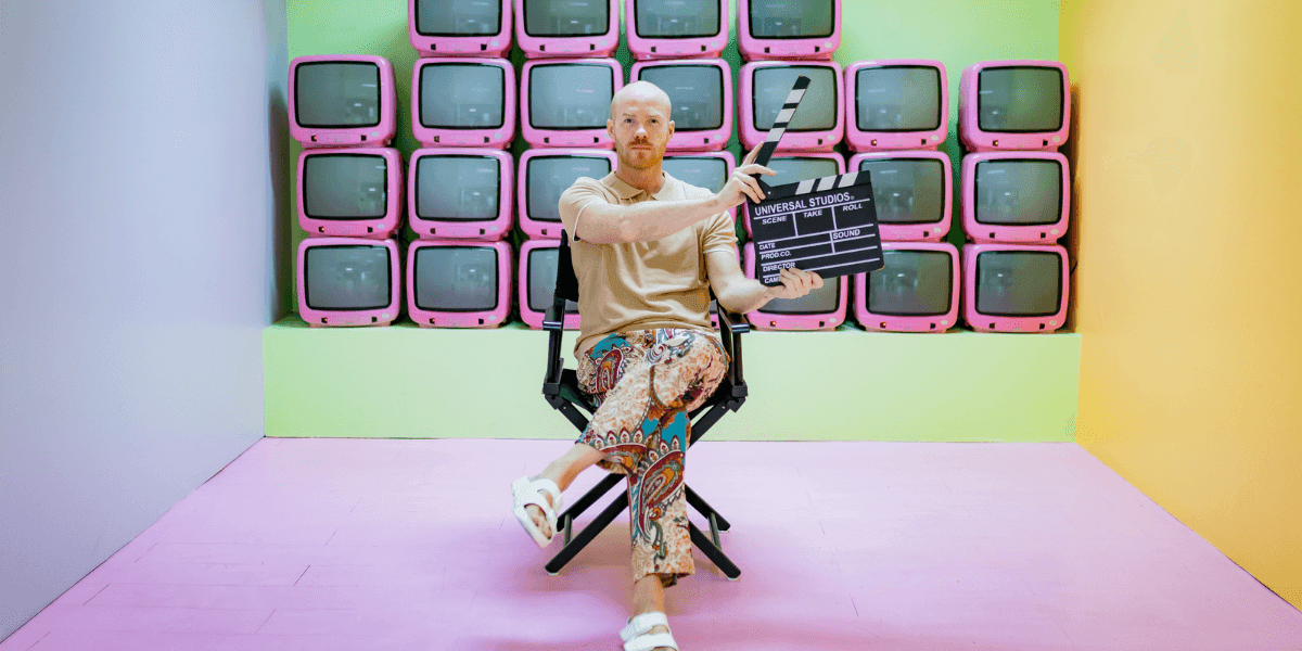 Thom Rikko holds a film clapperboard while seated before a colorful wall of vintage televisions.