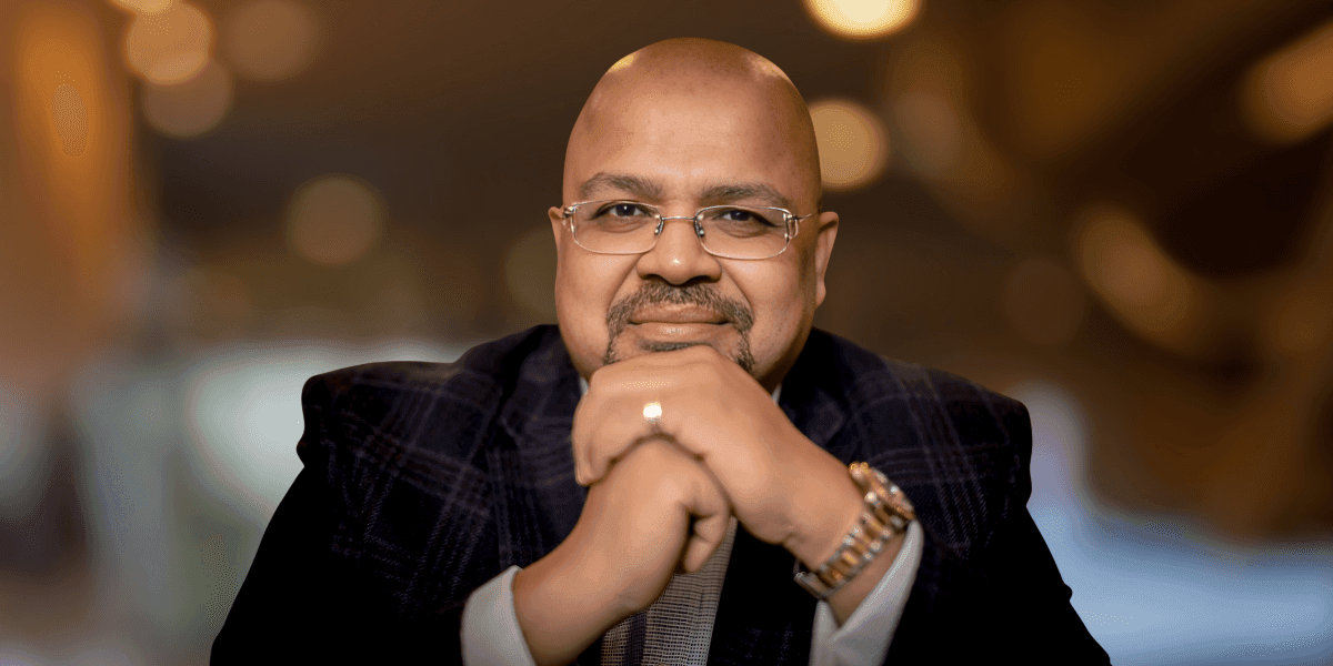 Man in glasses and suit jacket smiling with hands clasped under chin.