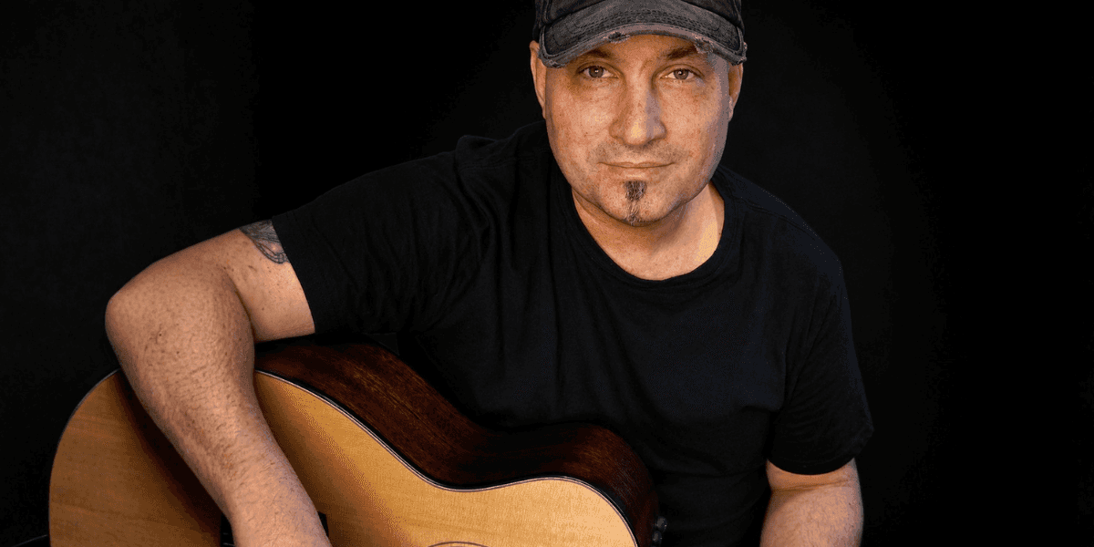 Man wearing black shirt and cap holding acoustic guitar against dark background.