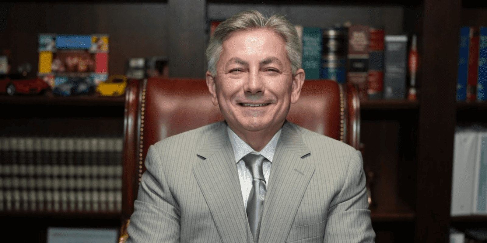 "Smiling man in a light gray suit and silver tie, sitting confidently in a leather chair with a bookshelf filled with legal books and collectible cars in the background."