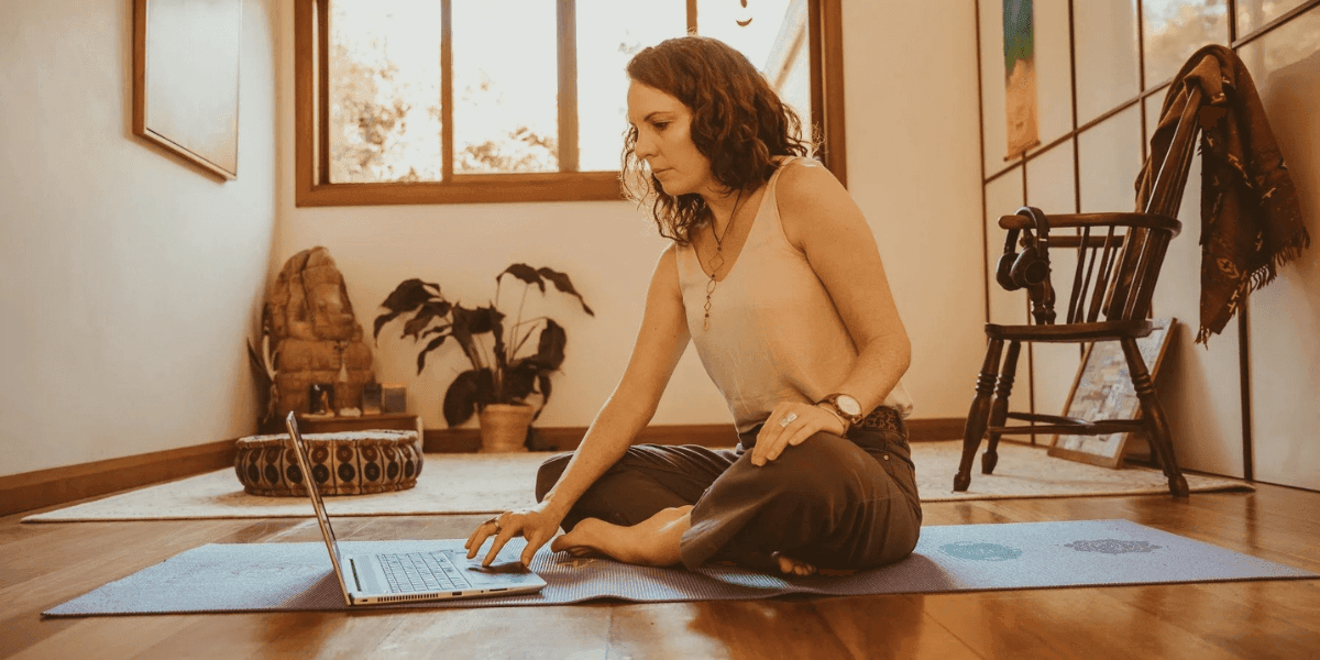 Woman sitting on a yoga mat using a laptop in a bright home wellness space.