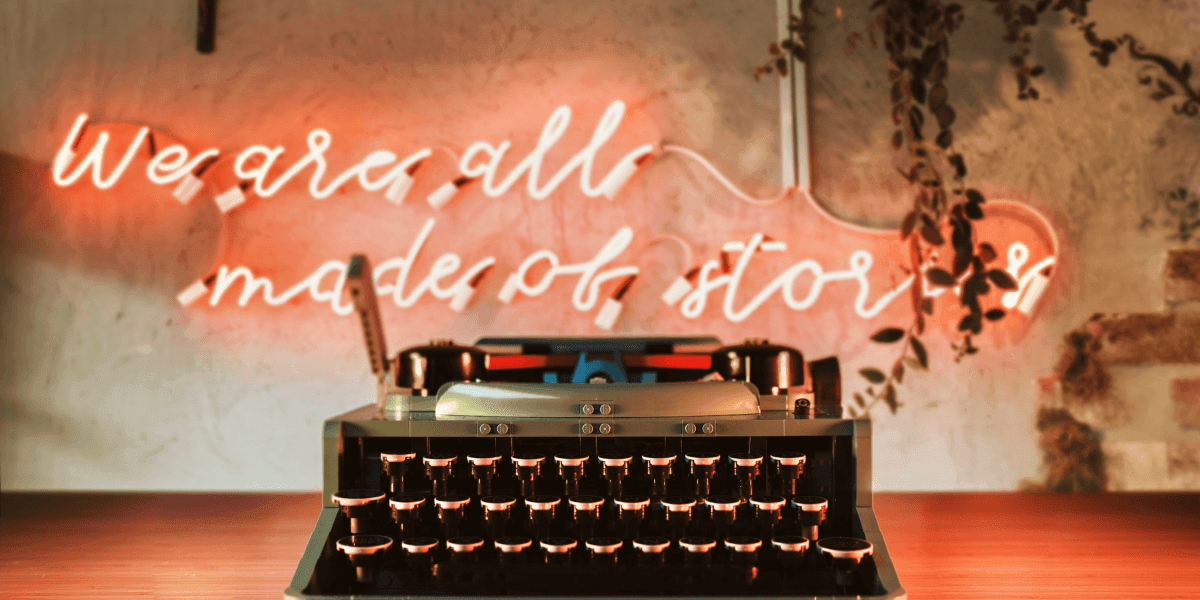 The image features a typewriter with a neon sign in the background reading, "We are all made of stories."