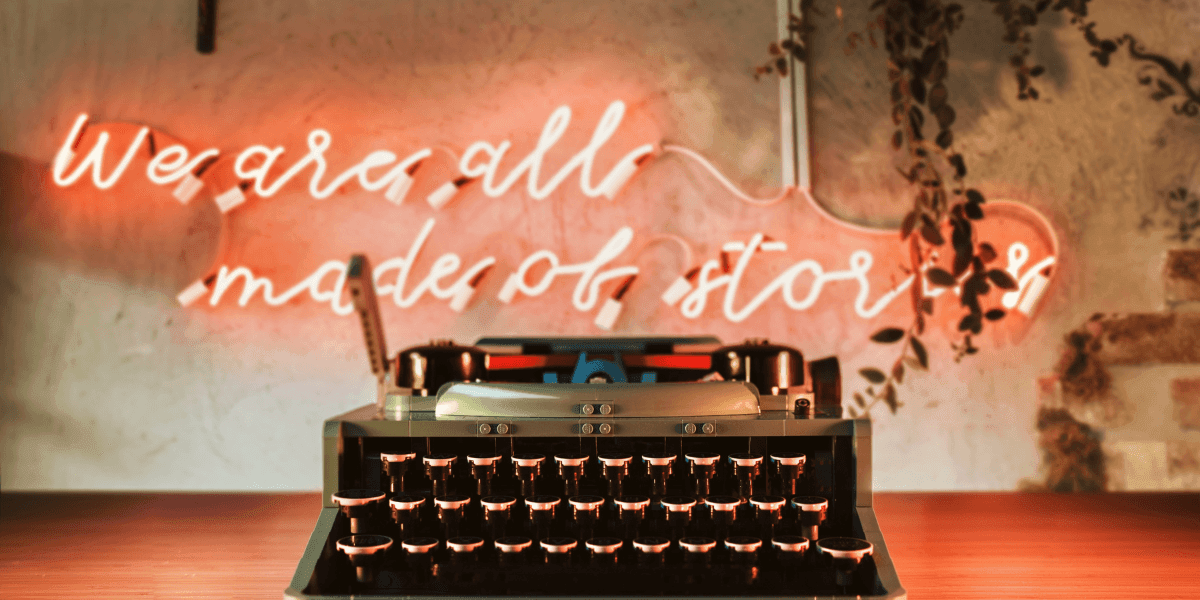 The image features a typewriter with a neon sign in the background reading, "We are all made of stories."