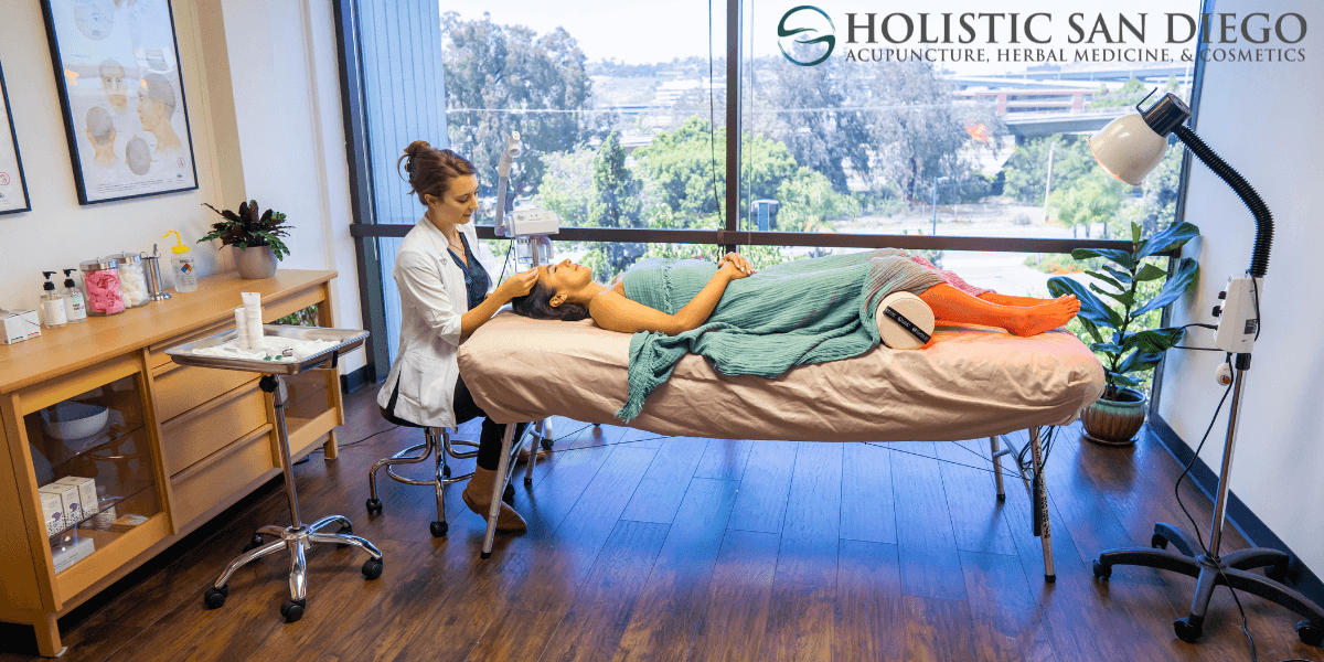 Holistic San Diego wellness clinic interior showing an esthetician performing a facial treatment on a relaxed female client. The treatment room features a large window with a scenic view, modern medical spa equipment, skincare products, and a peaceful ambiance. Focus on holistic health, acupuncture, herbal medicine, and cosmetic care.