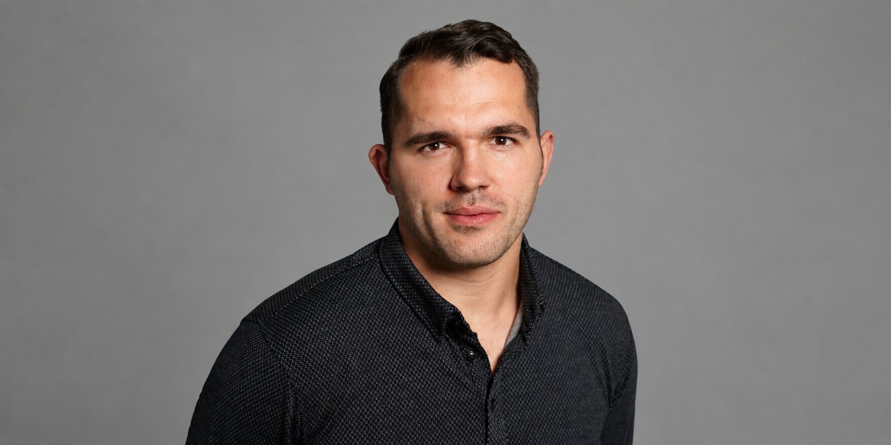 Professional headshot of Tim Tesluck wearing a black button-down shirt against a gray background.