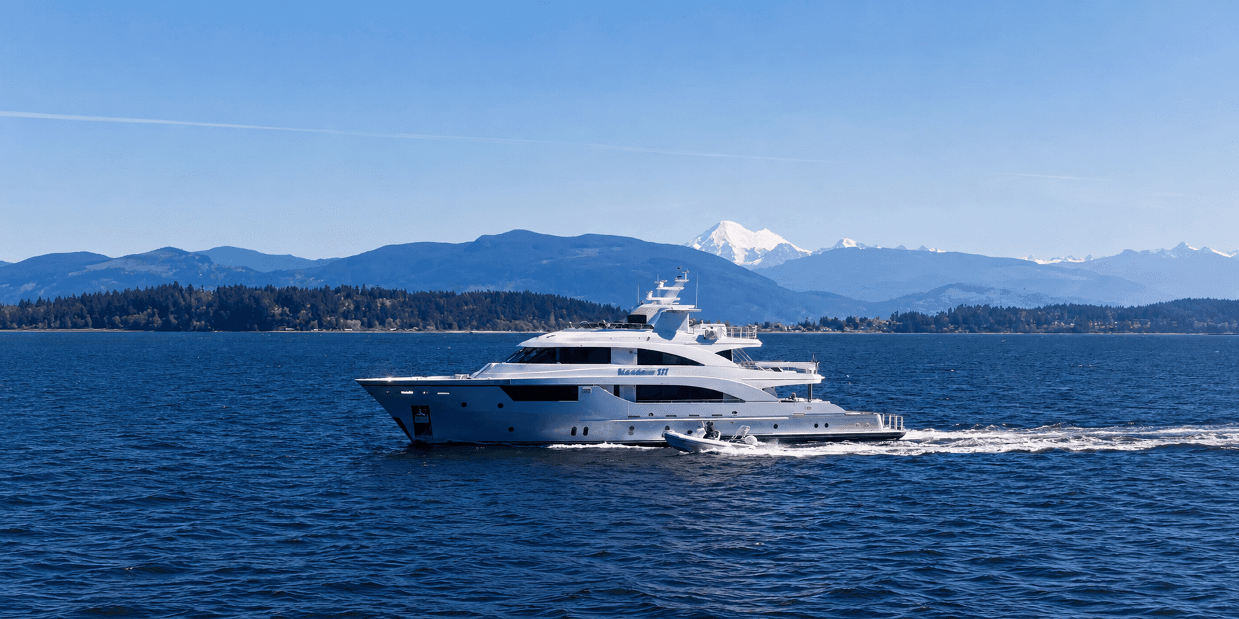 MAXIMUS III cruising across blue water with forested hills and snowcapped mountains in the background.