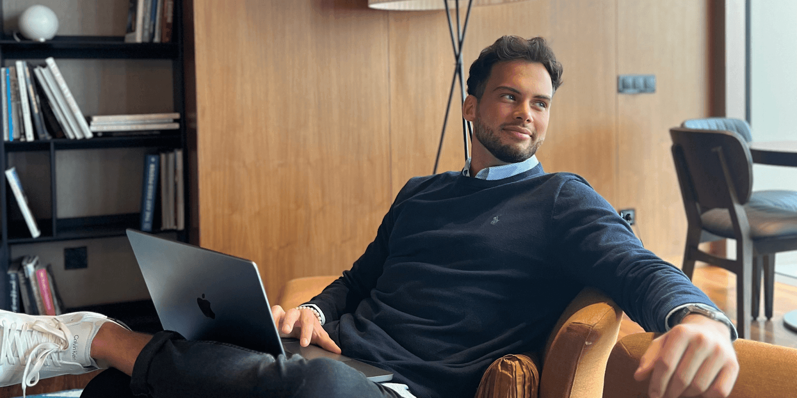 Man sitting in a chair with a laptop, looking to the side in a modern, wood-paneled room.