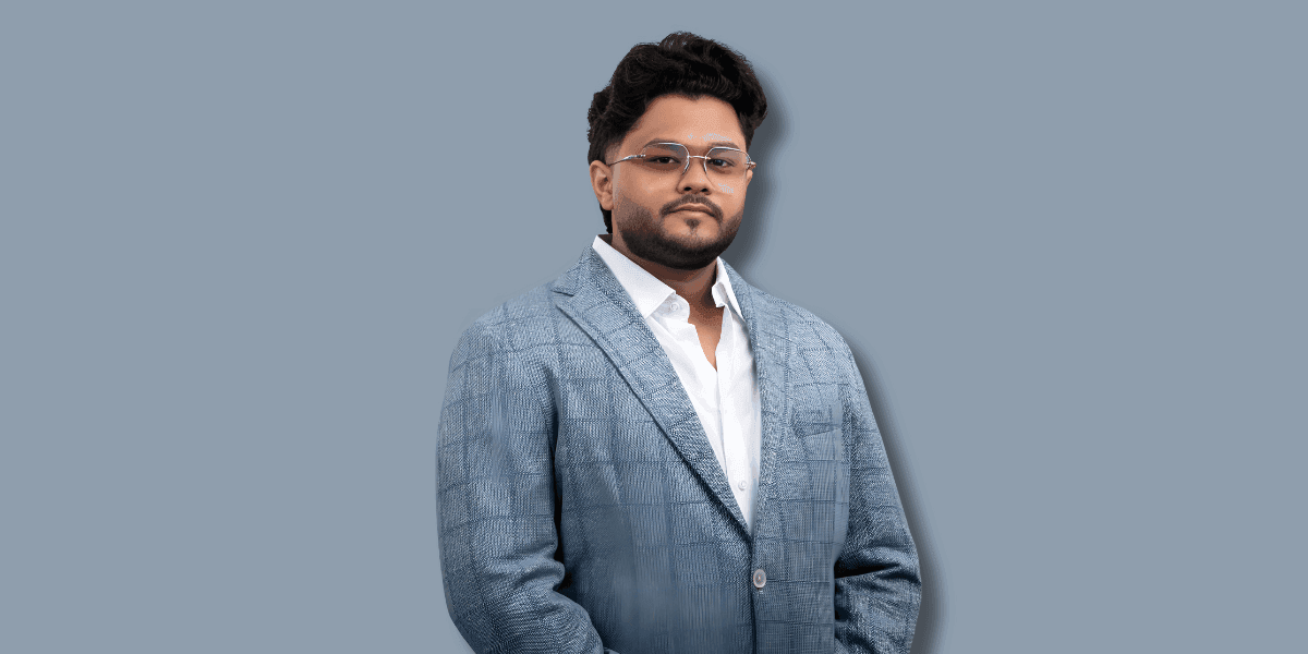 Man in blue checkered suit and glasses posing confidently against a plain grey background.