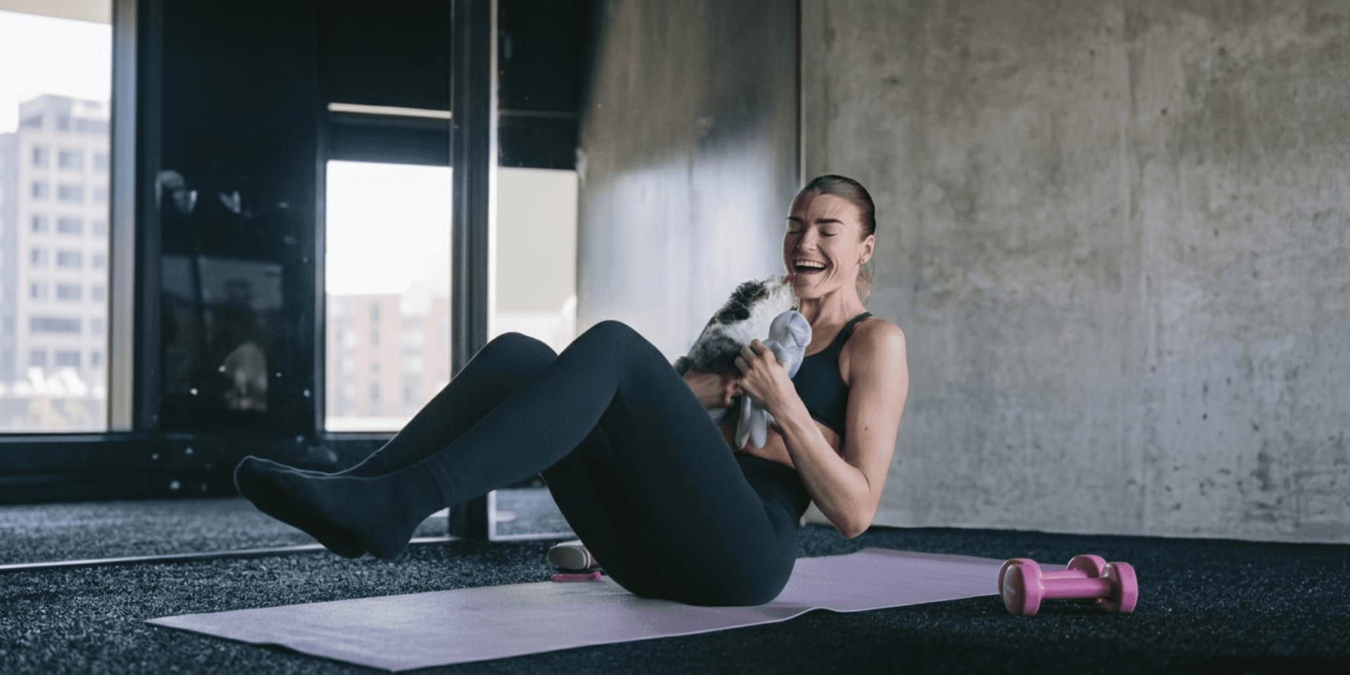 Woman doing Pilates on a mat while smiling and holding a playful puppy indoors.