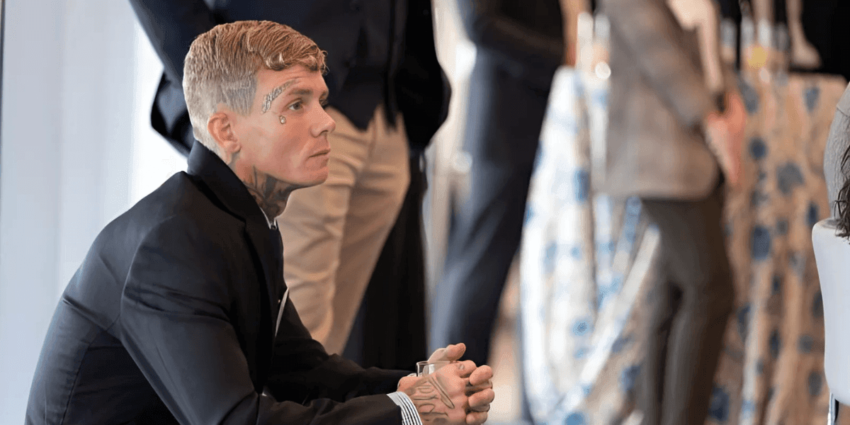 Man with face tattoos in formal attire sits thoughtfully at networking event, symbolizing personal transformation and leadership.