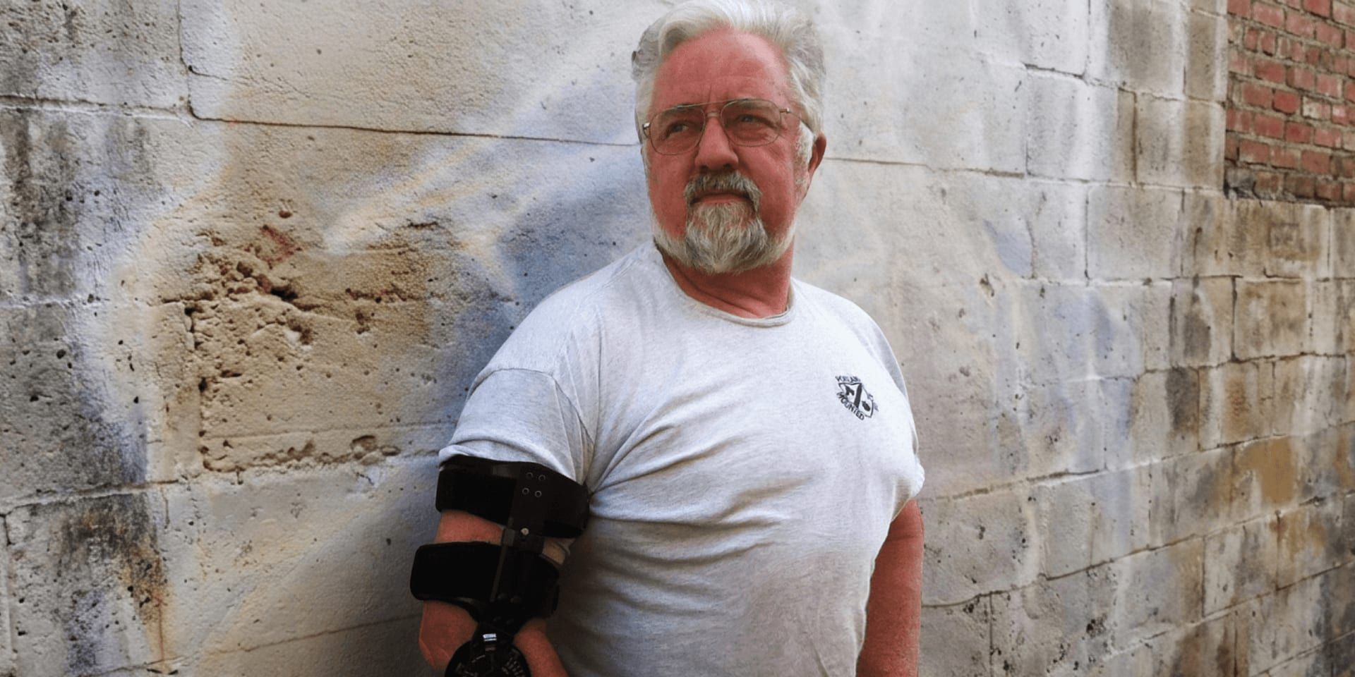 Baer Charlton standing outdoors beside a weathered wall wearing an arm brace.