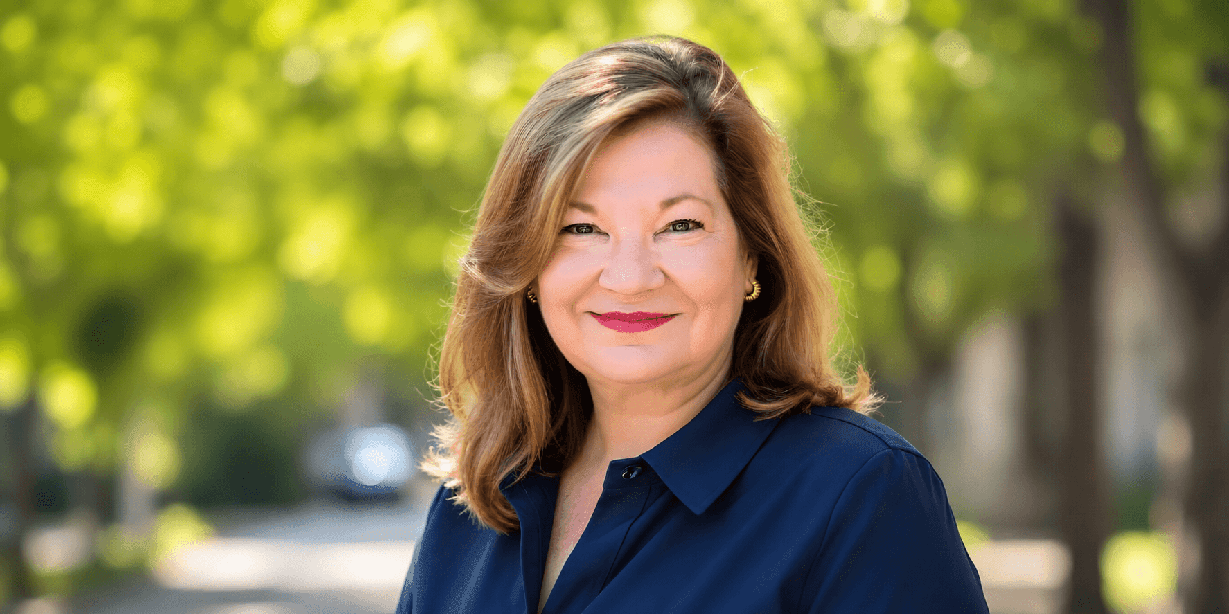 Professional headshot of a smiling female executive outdoors, wearing a navy blouse with a blurred green background.