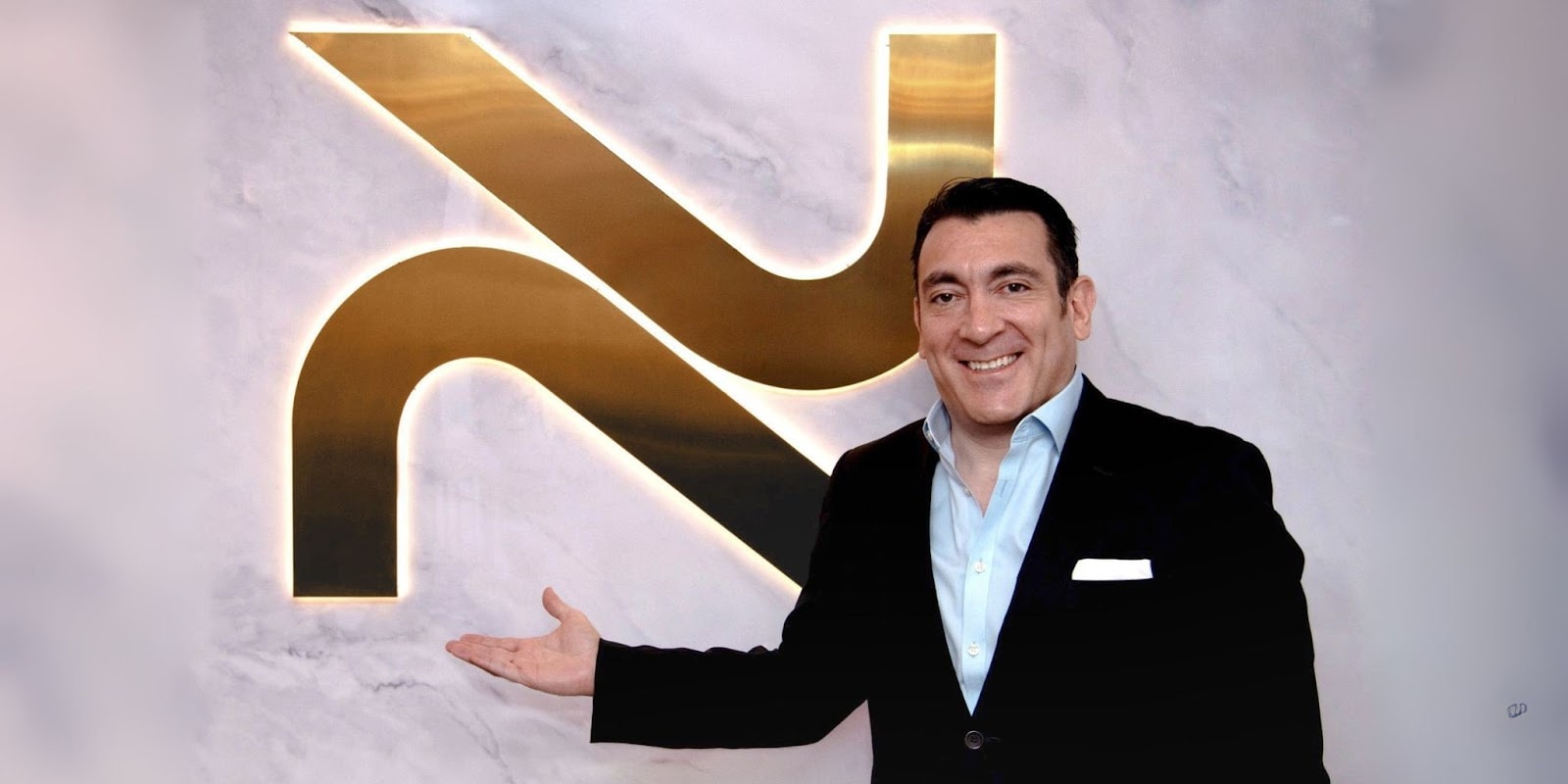 A man standing next to a glowing NovoSculpt logo on a marble wall, smiling and presenting the brand with a welcoming gesture. The logo represents the NovoSculpt fat removal technology.