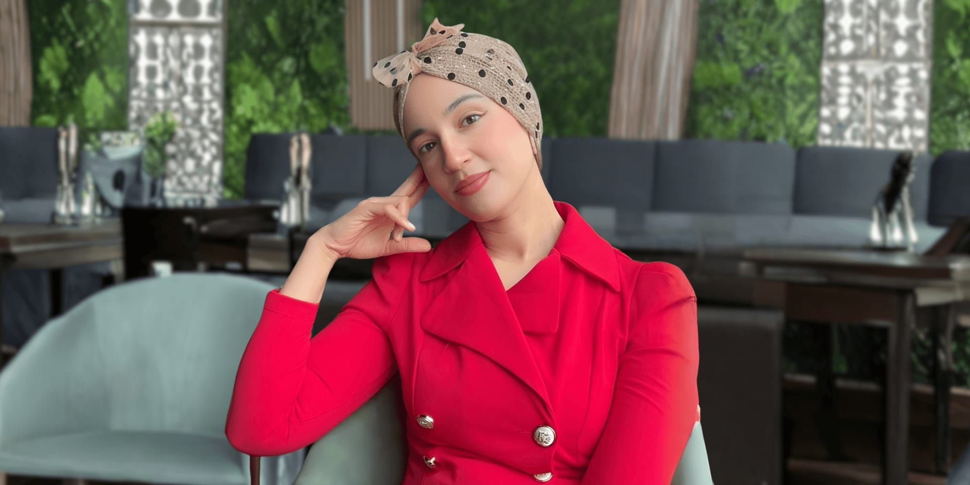 Woman in red blazer and polka-dot headscarf sitting thoughtfully in a modern café setting.
