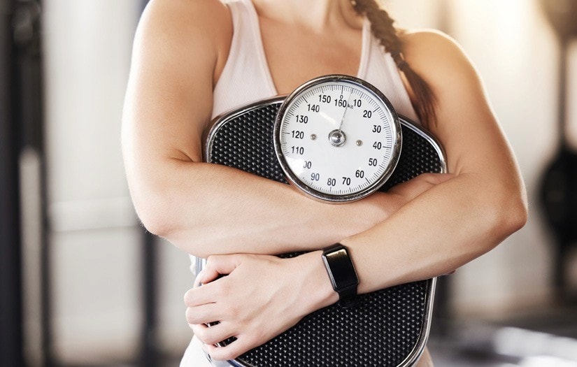 Person in workout attire hugging bathroom scale, symbolizing weight loss and body composition focus.