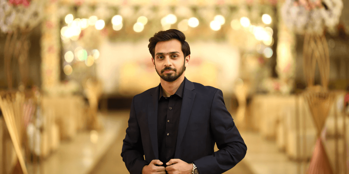 "A professional young man dressed in a black suit, standing confidently at a luxury event venue with a blurred background of soft lighting and elegant decor."