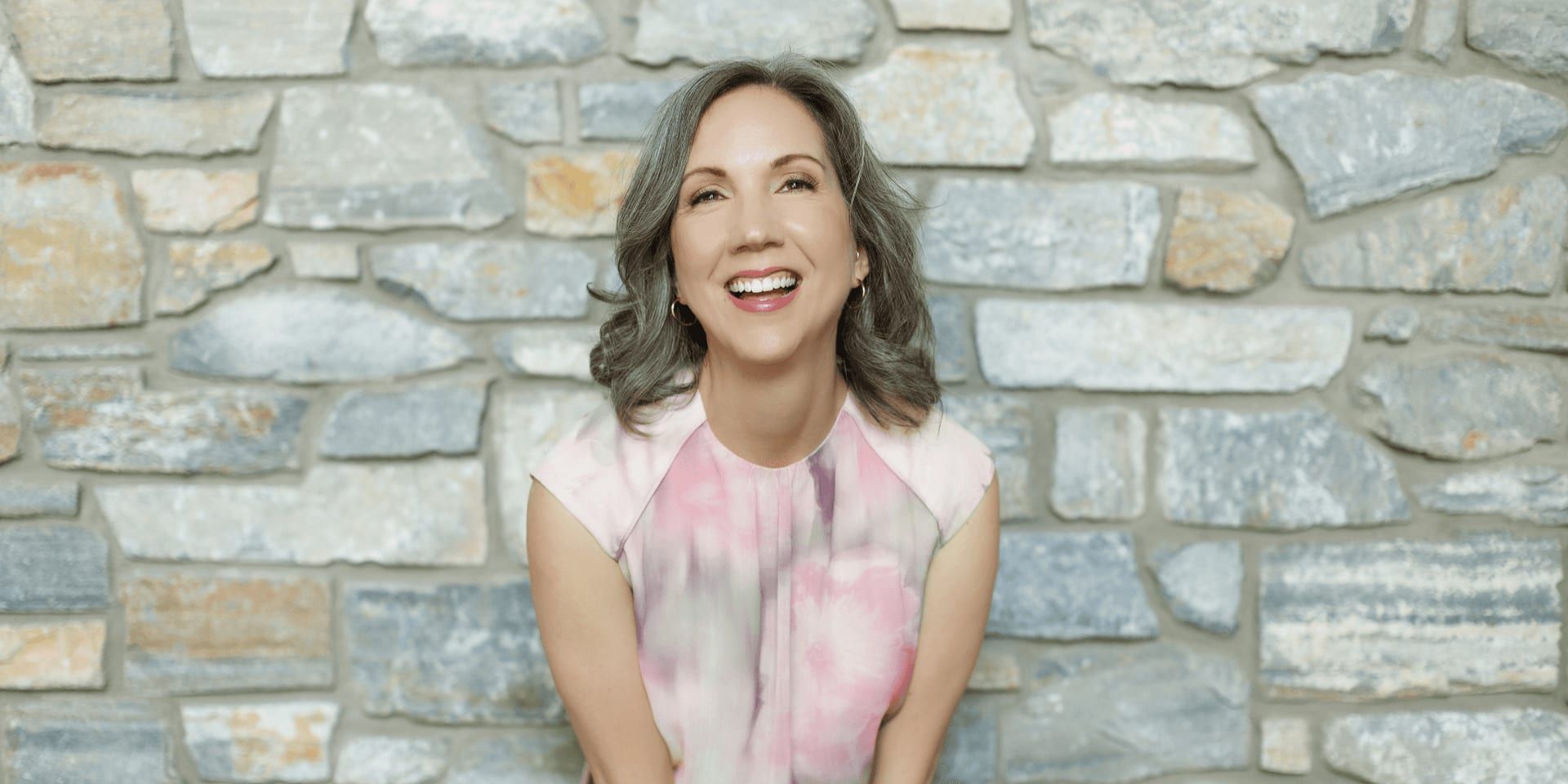 Smiling woman in pink blouse standing before a stone wall background, conveying warmth and confidence.