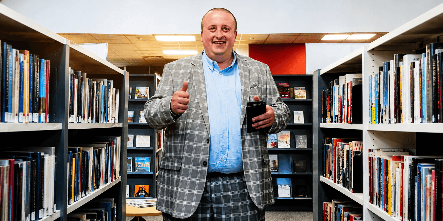 Jawad Sharief standing between library bookshelves holding a phone and giving a thumbs-up.