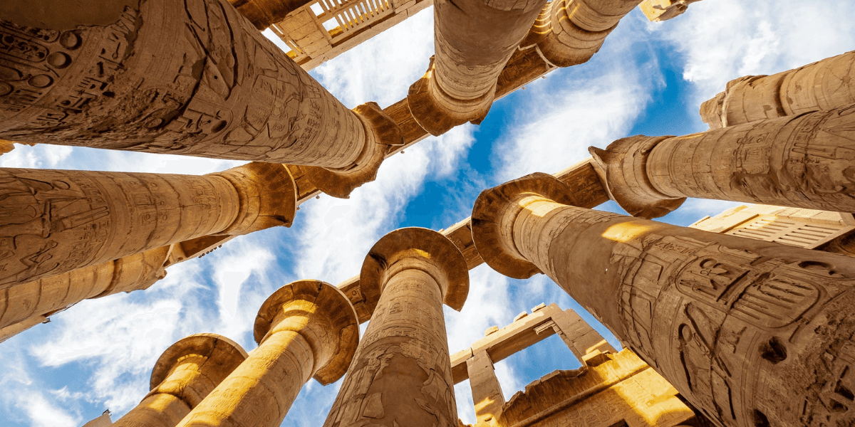 Upward view of ancient Egyptian temple columns with carved hieroglyphs against a blue sky.